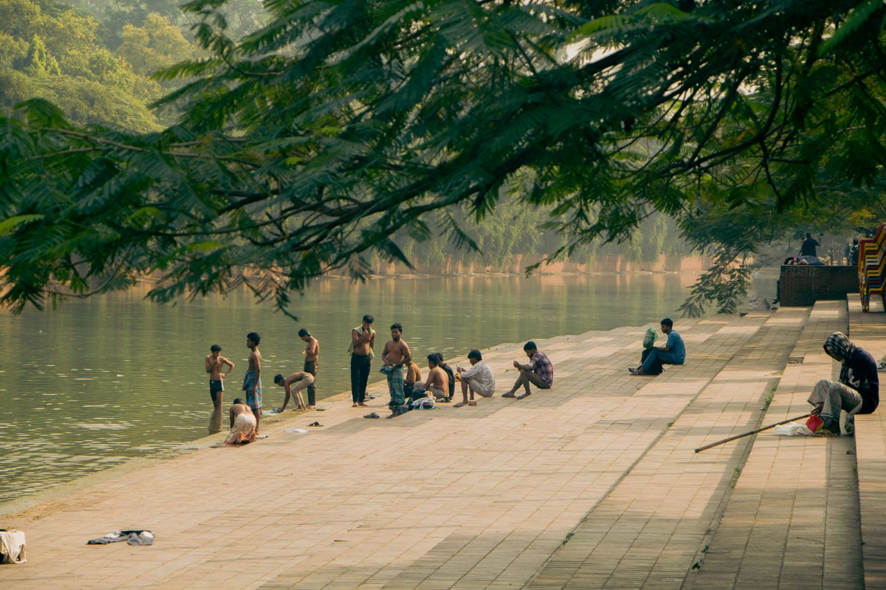A serene scene of people enjoying the riverside in Dhaka, Bangladesh, under lush greenery.